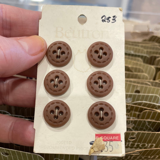 Hand holding a card with six brown buttons against a blurred background of more buttons and containers.