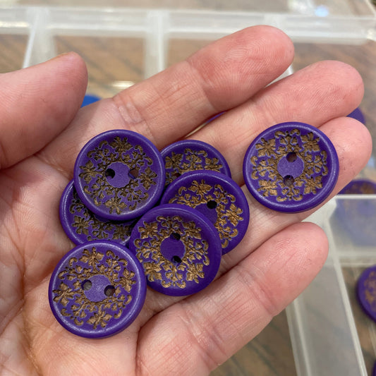 Hand holding purple buttons with gold patterns in front of a clear container.