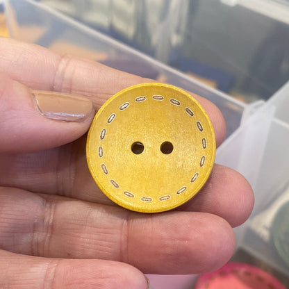 Hand holding a yellow button with two holes, with a container of buttons in the background.