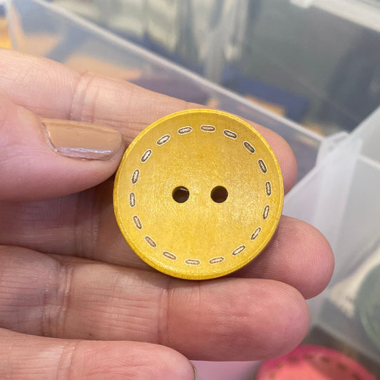 Hand holding a yellow button with two holes, with a container of buttons in the background.