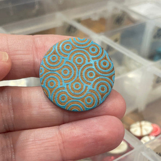 Hand holding a decorative button with a pattern, with a container of buttons in the background.