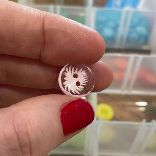 Hand holding a small round object with a design, against a background of a clear plastic container with compartments.