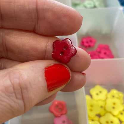 Hand holding two small flower-shaped earrings with a container of more earrings in the background.
