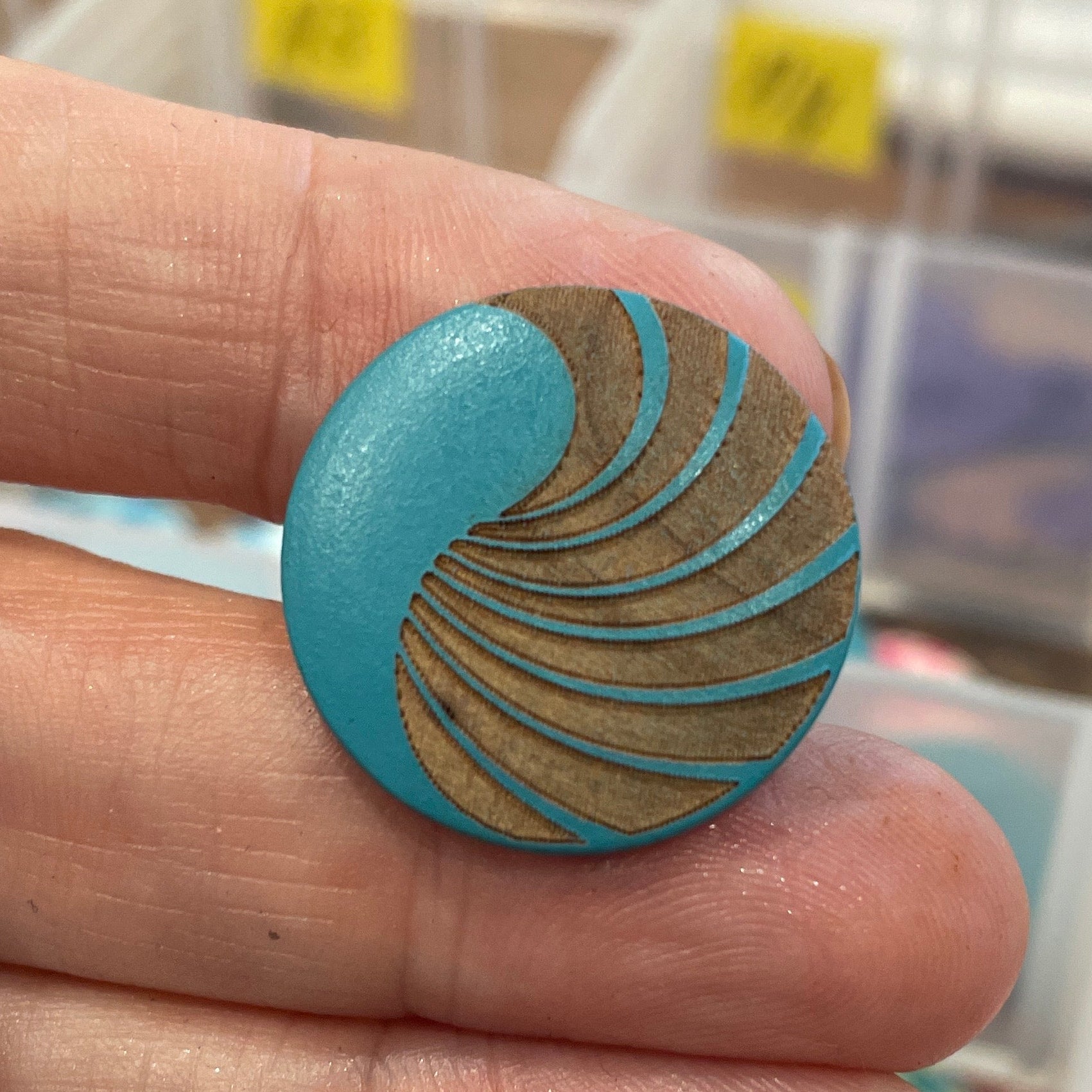 Close-up of a hand holding a blue and gold button with a blurred background of button containers.