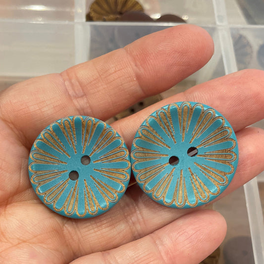 Hand holding two blue buttons with gold patterns against a background of more buttons in a container.