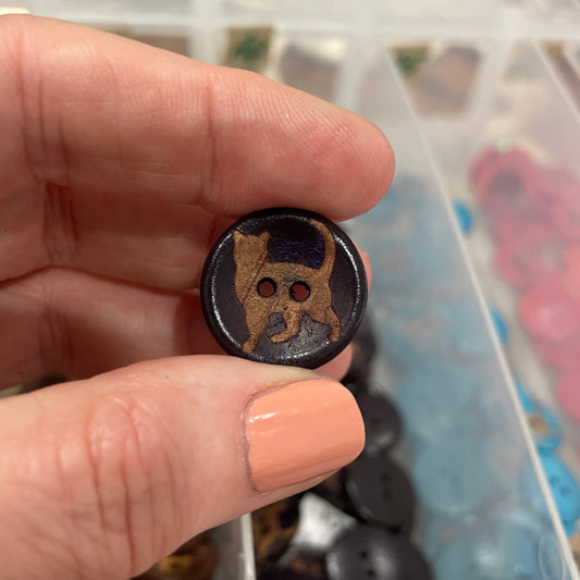 Hand holding a black button with two holes in front of a container of buttons.