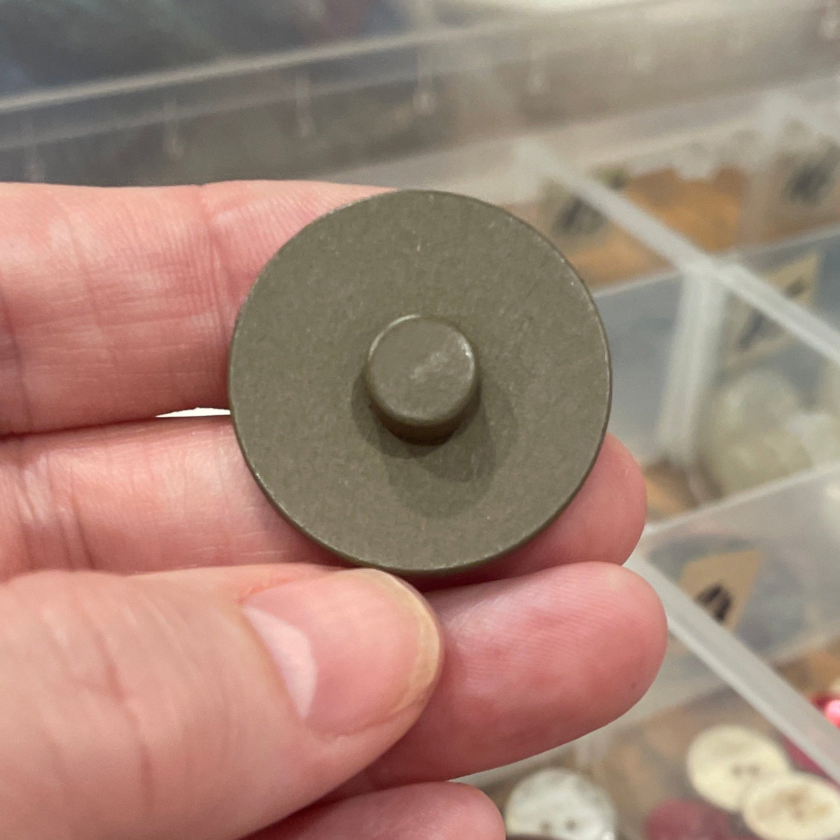 Hand holding a round metal component with a background of a divided storage container filled with various items.