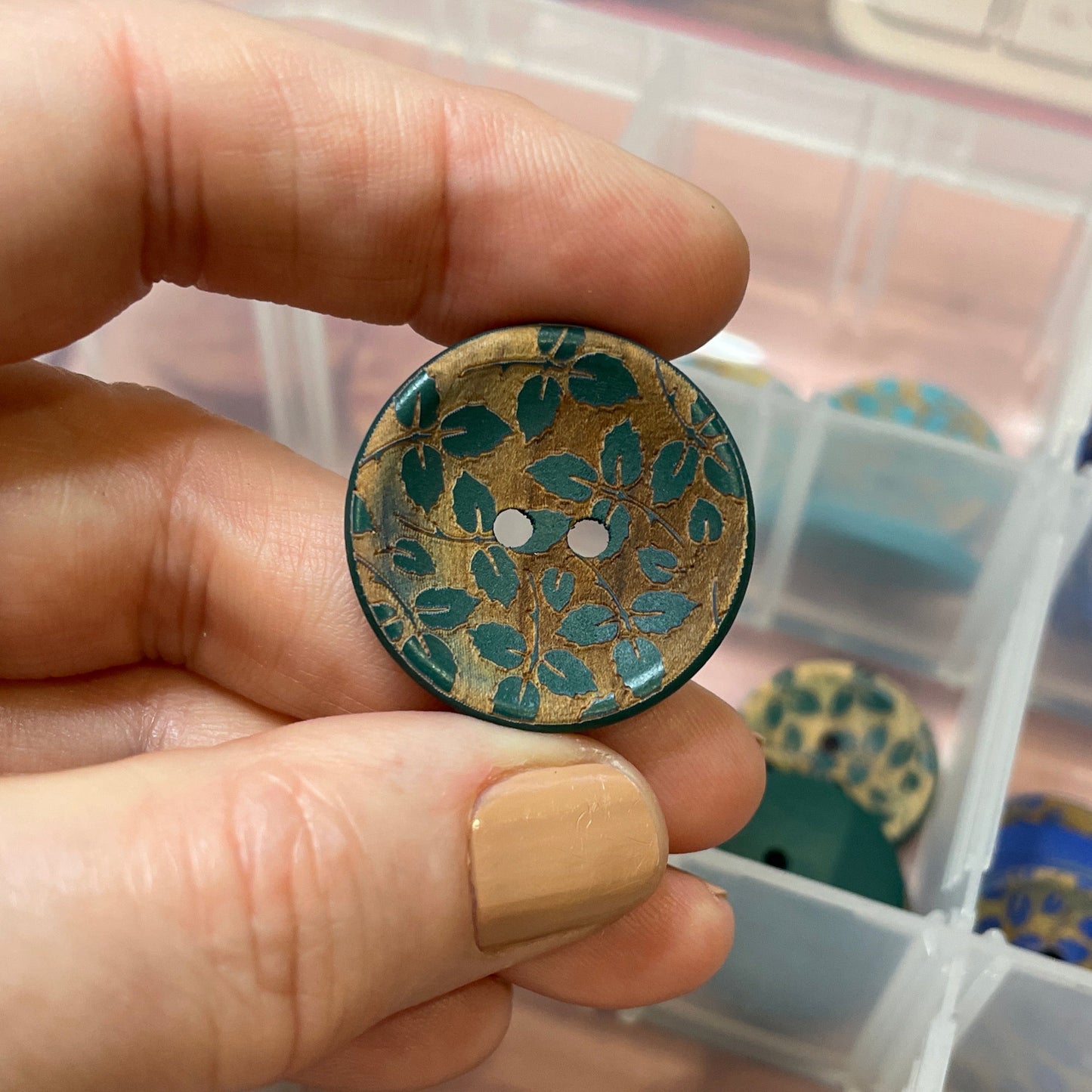 Hand holding a decorative button with a container of various buttons in the background