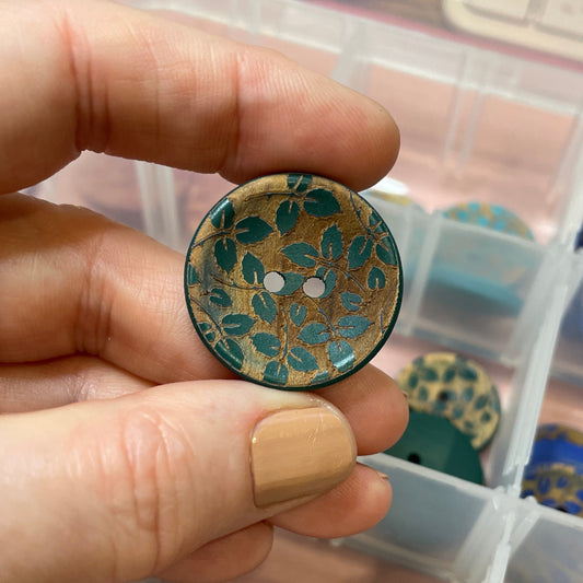 Hand holding a decorative button with a container of various buttons in the background