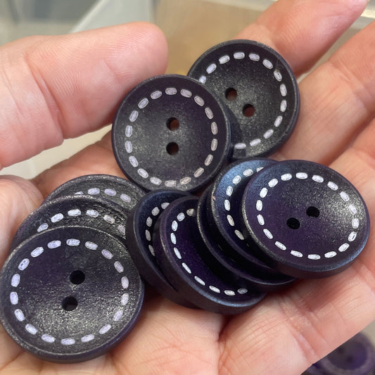 Hand holding black buttons with white stitching, blurred container of buttons in the background