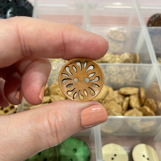 Hand holding a decorative wooden button in front of a container with various buttons.