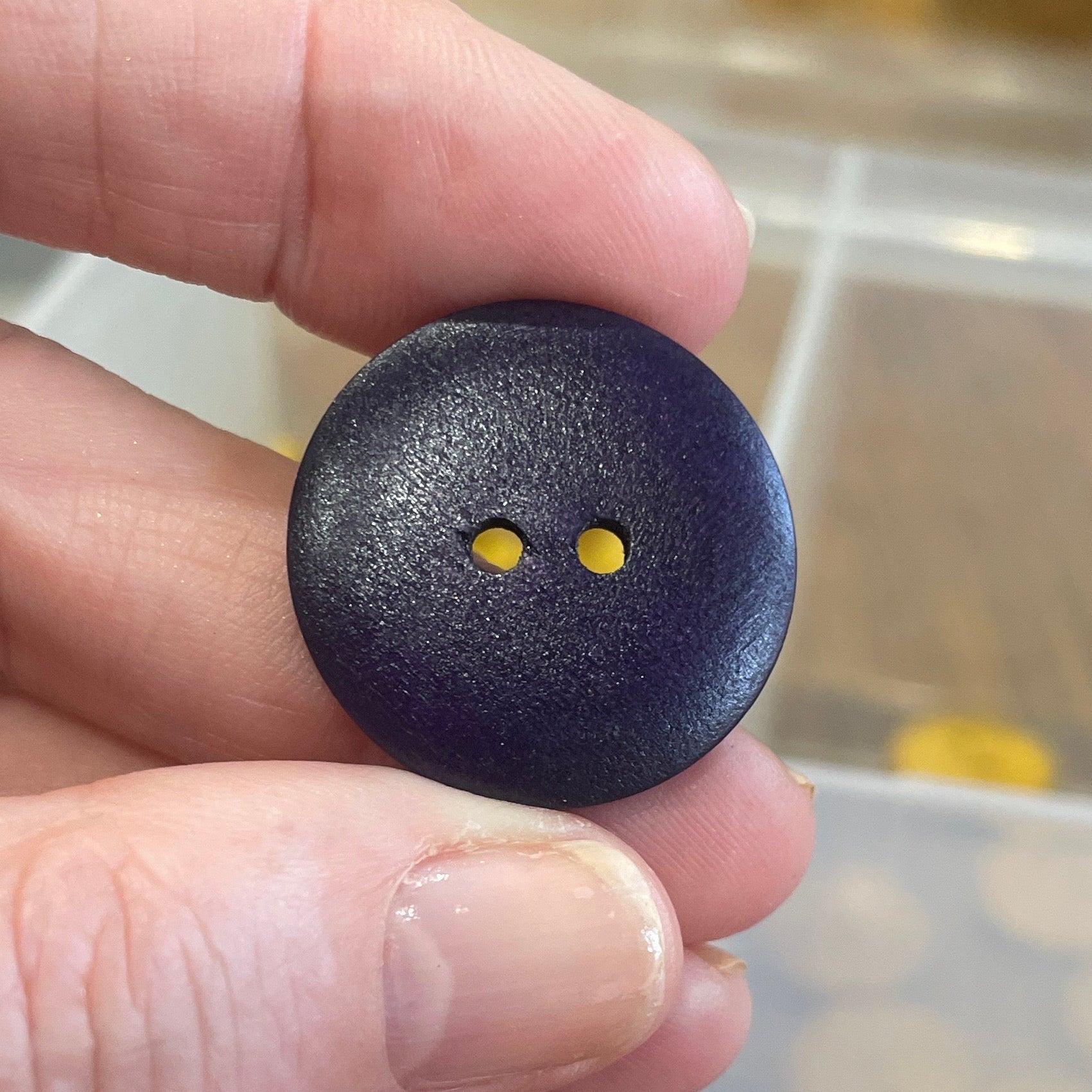 Hand holding a blue button with a blurred container of buttons in the background