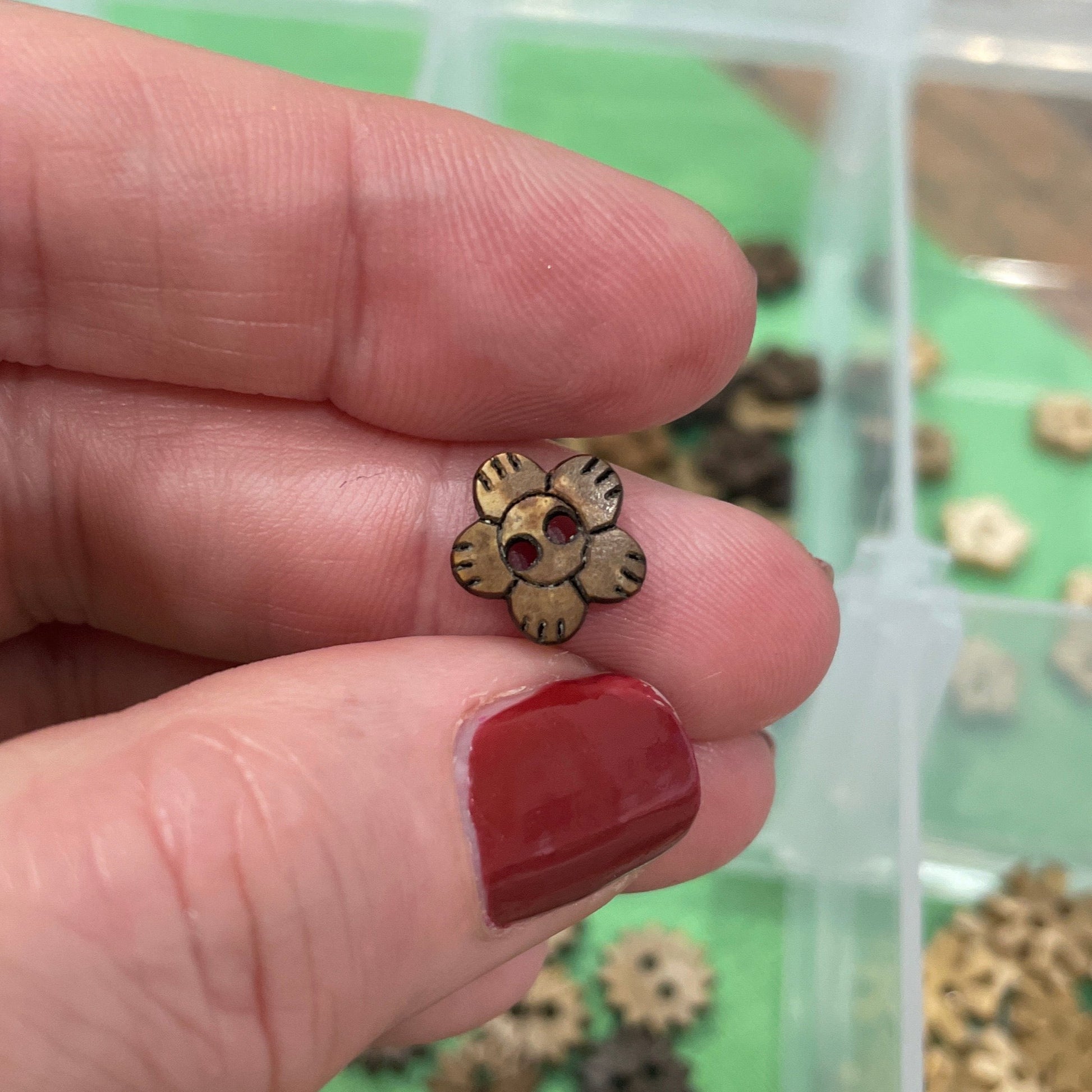 Hand holding a small gold flower-shaped button with a container of similar objects in the background.