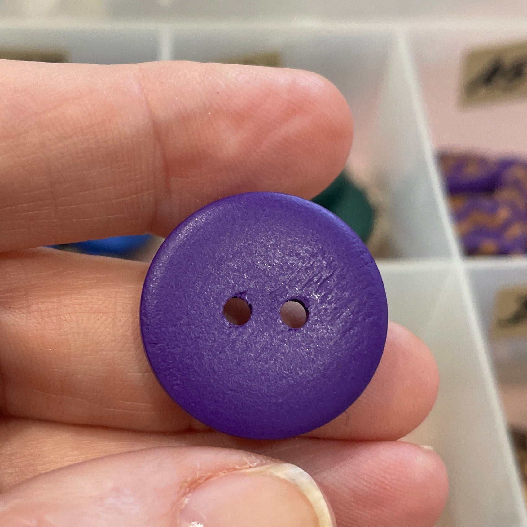 Purple button held between fingers with a blurred background of a storage box with buttons.