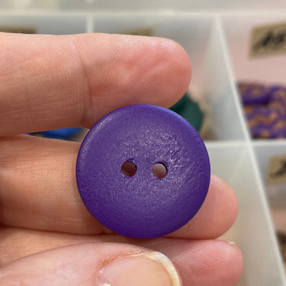 Purple button held between fingers with a blurred background of a storage box with buttons.