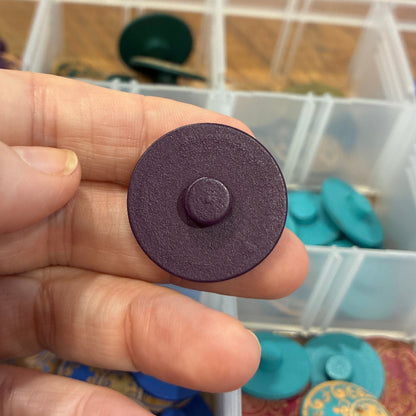 Hand holding a round purple button with a background of button containers.