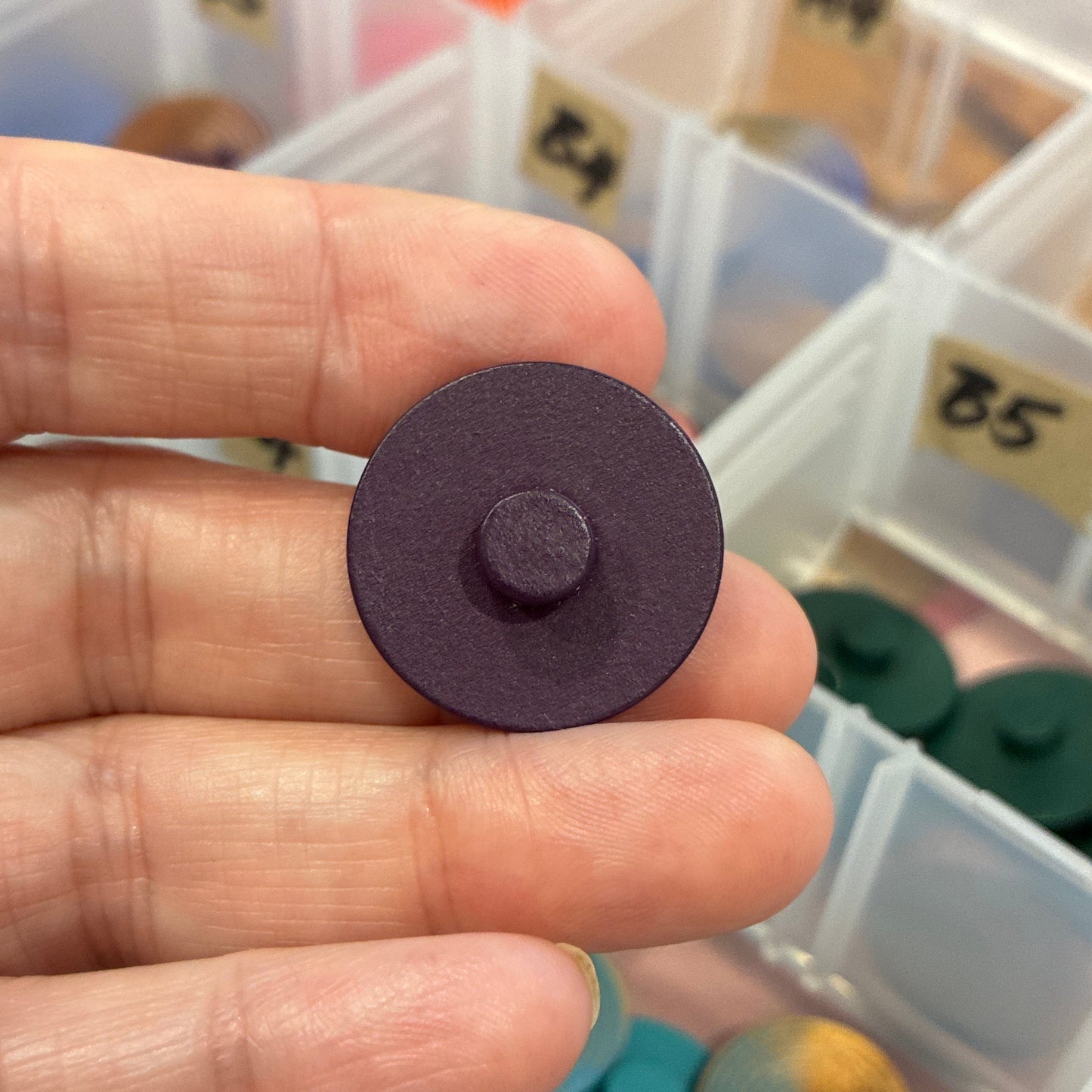 Hand holding a purple button with a container of buttons in the background