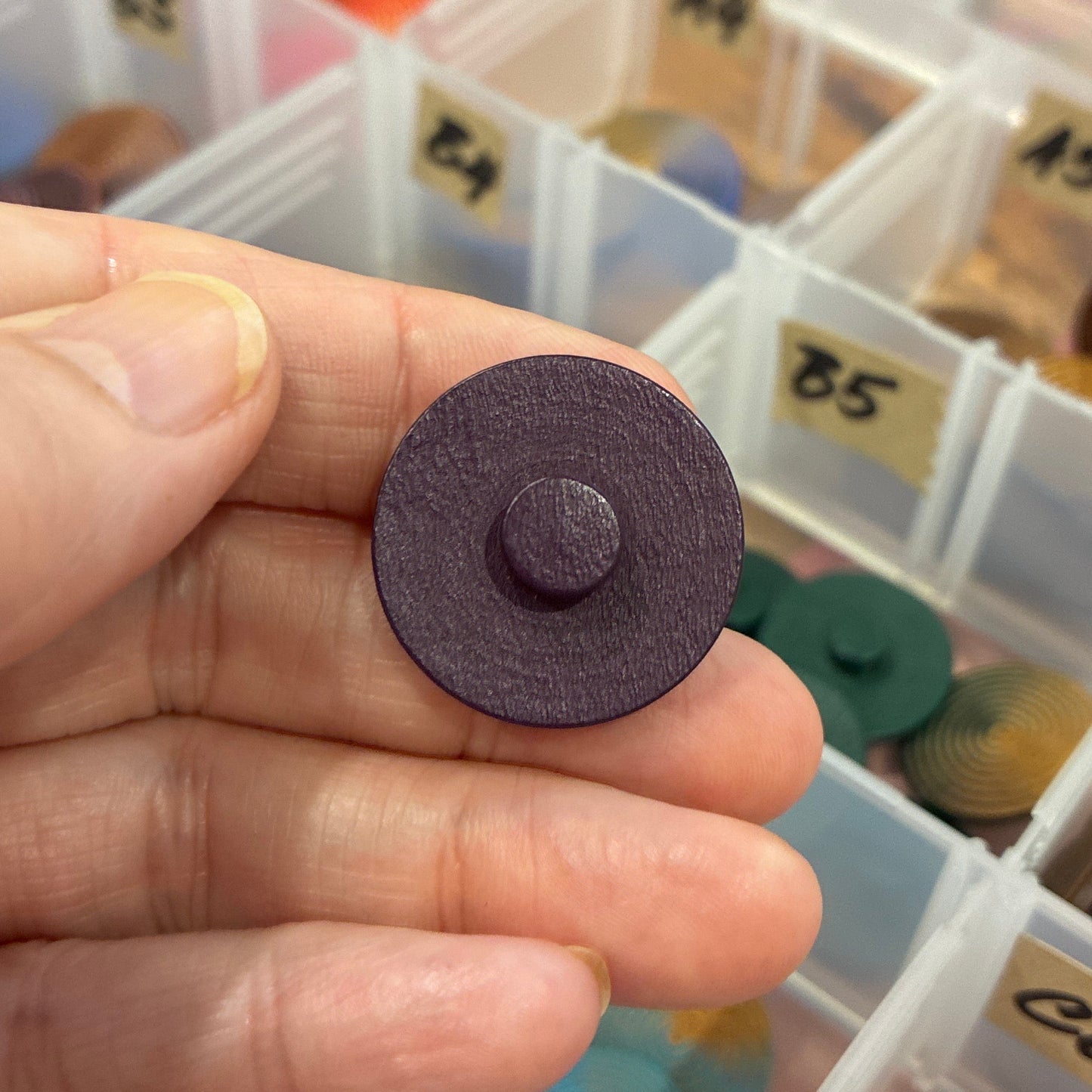 Hand holding a purple button with a container of buttons in the background