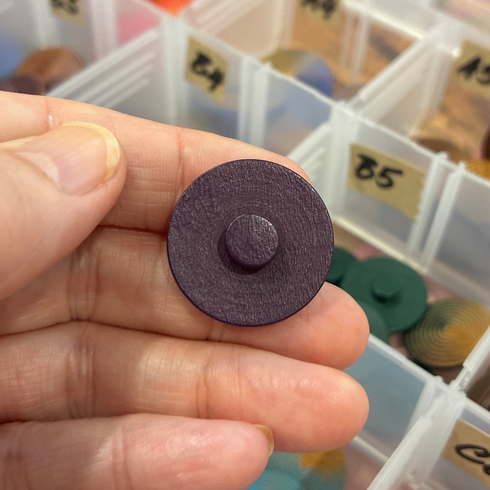 Hand holding a purple button with a container of buttons in the background