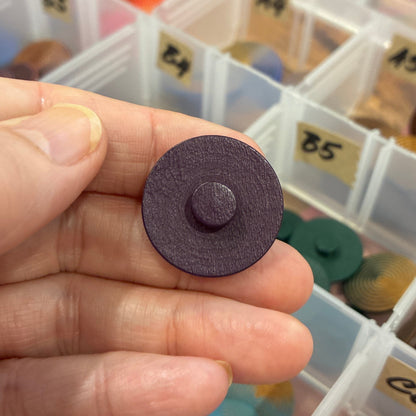 Hand holding a purple button with a container of buttons in the background