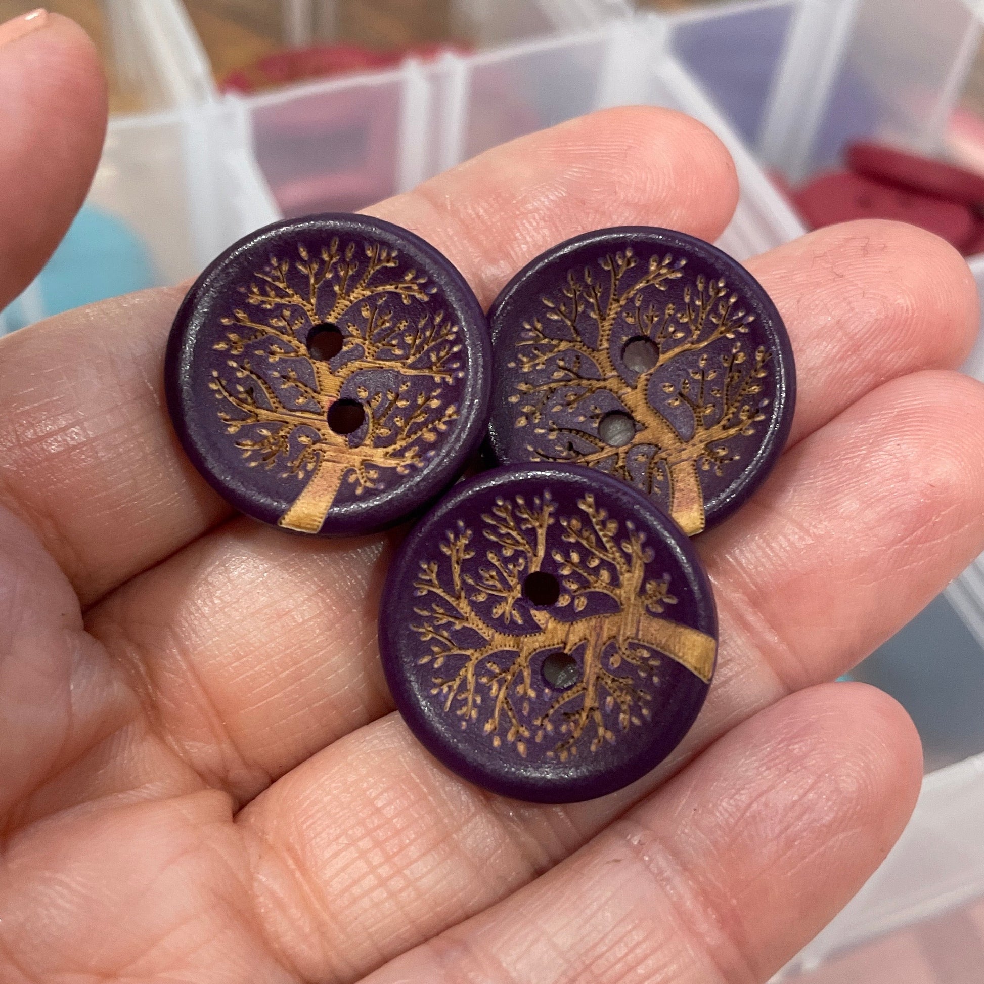 Hand holding three purple buttons with gold designs against a blurred background of a storage container.