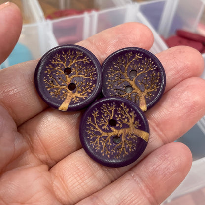 Hand holding three purple buttons with gold designs against a blurred background of a storage container.