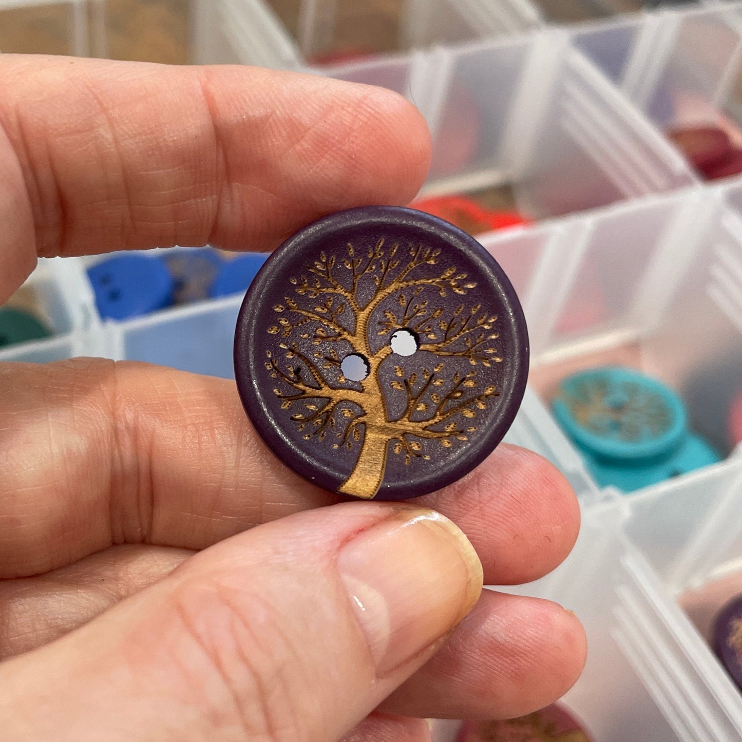 Hand holding a decorative button with intricate design, with a container of buttons in the background.