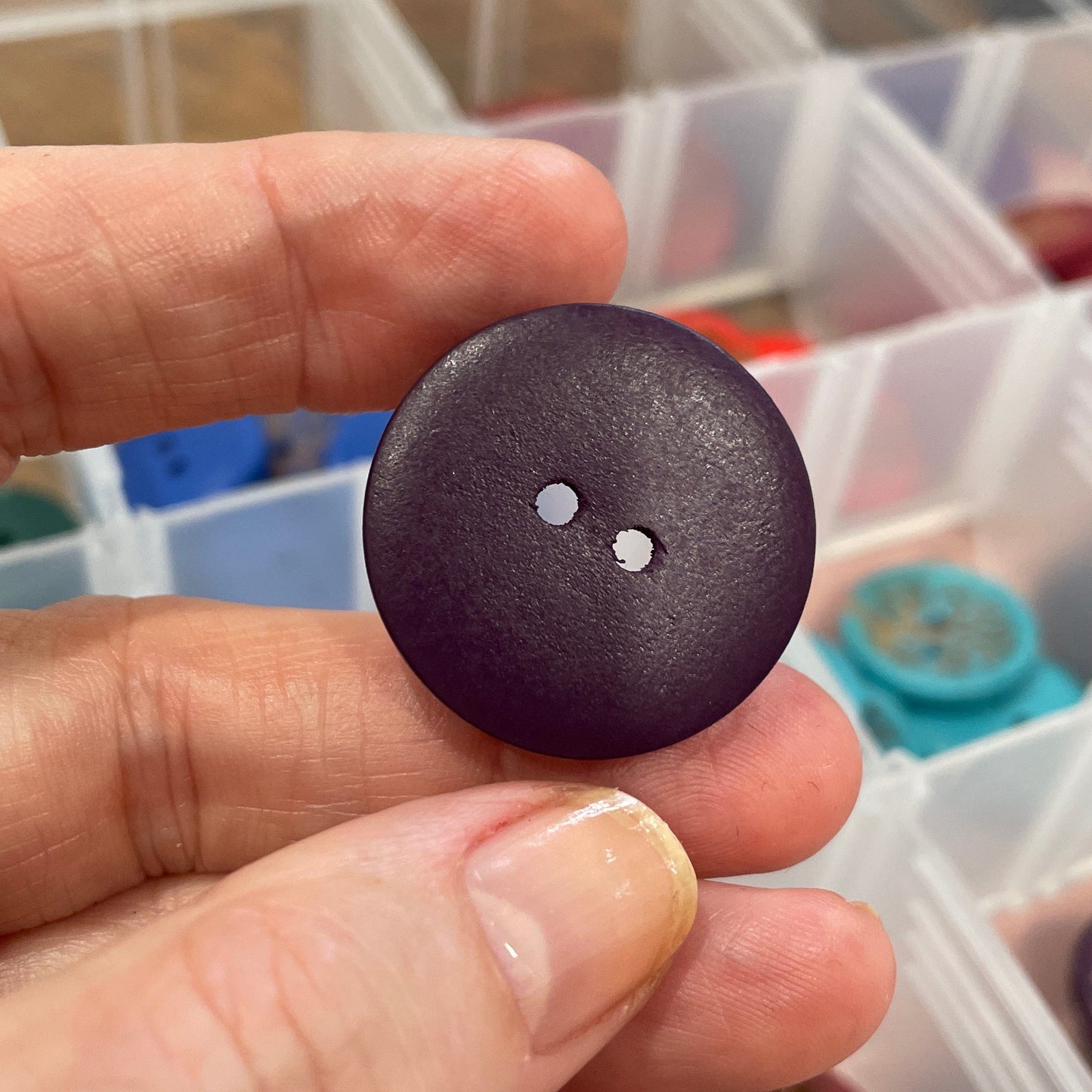 Hand holding a dark brown button with a clear container of buttons in the background