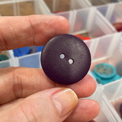 Hand holding a dark brown button with a clear container of buttons in the background