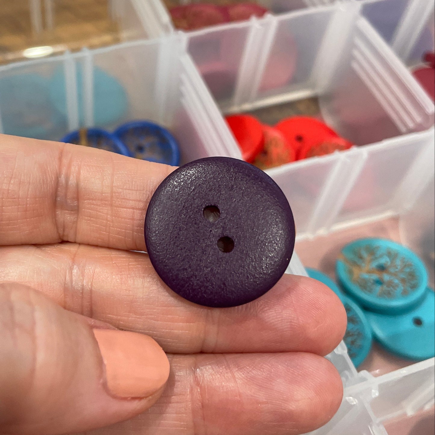 Hand holding a dark purple button with a background of colorful buttons in a container.