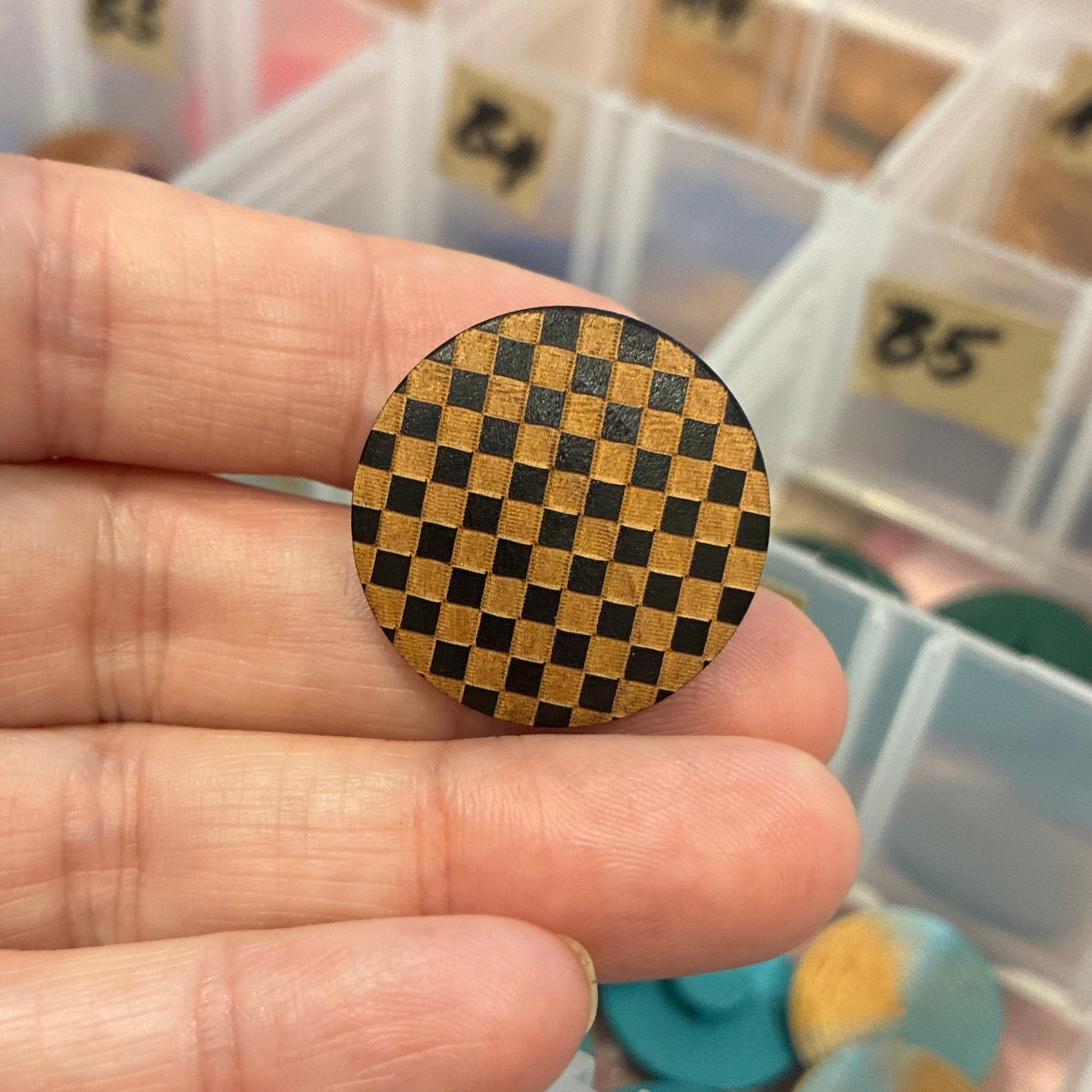 Hand holding a checkered round button with a transparent container of assorted items in the background