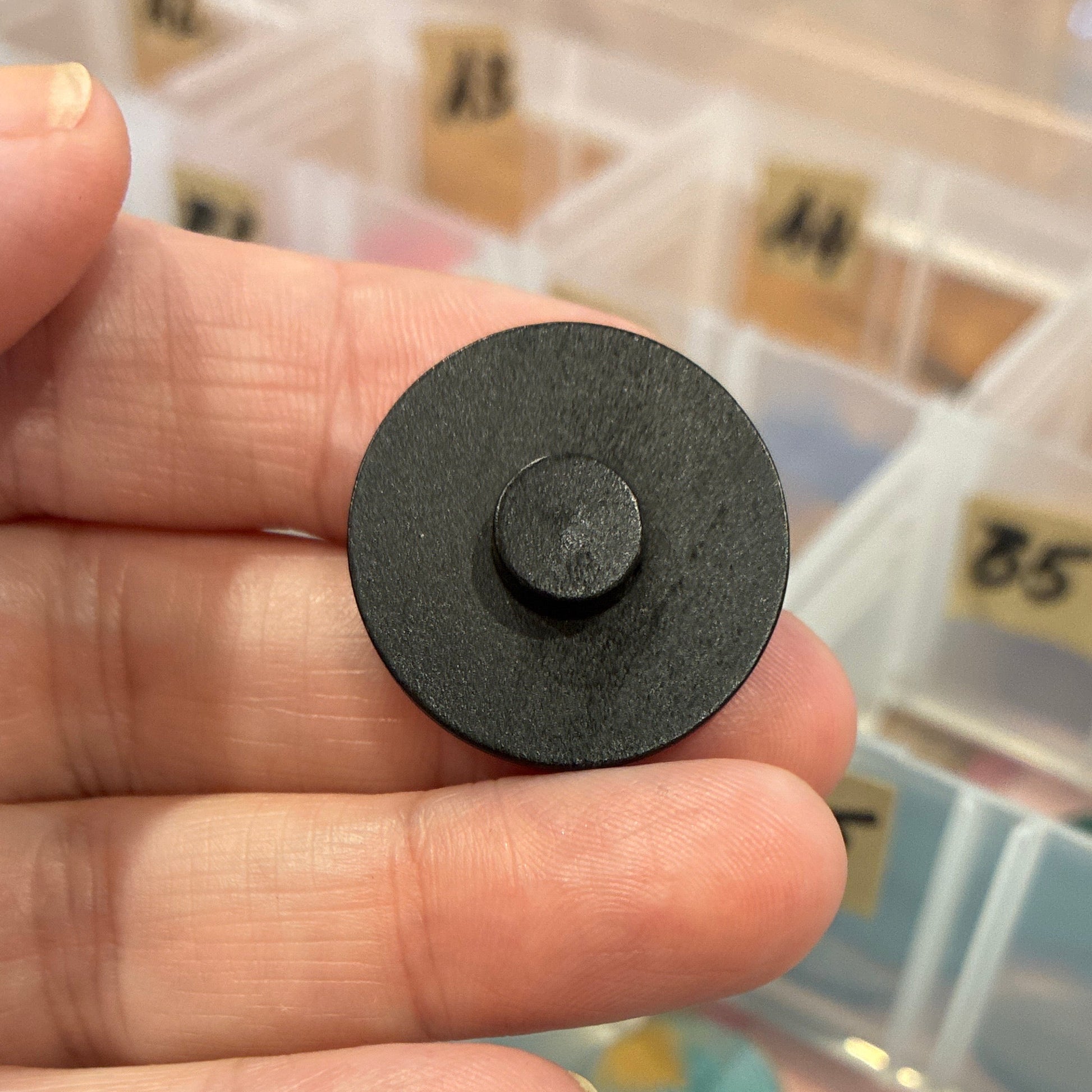 Black round button held by a hand with a container of assorted colored objects in the background.