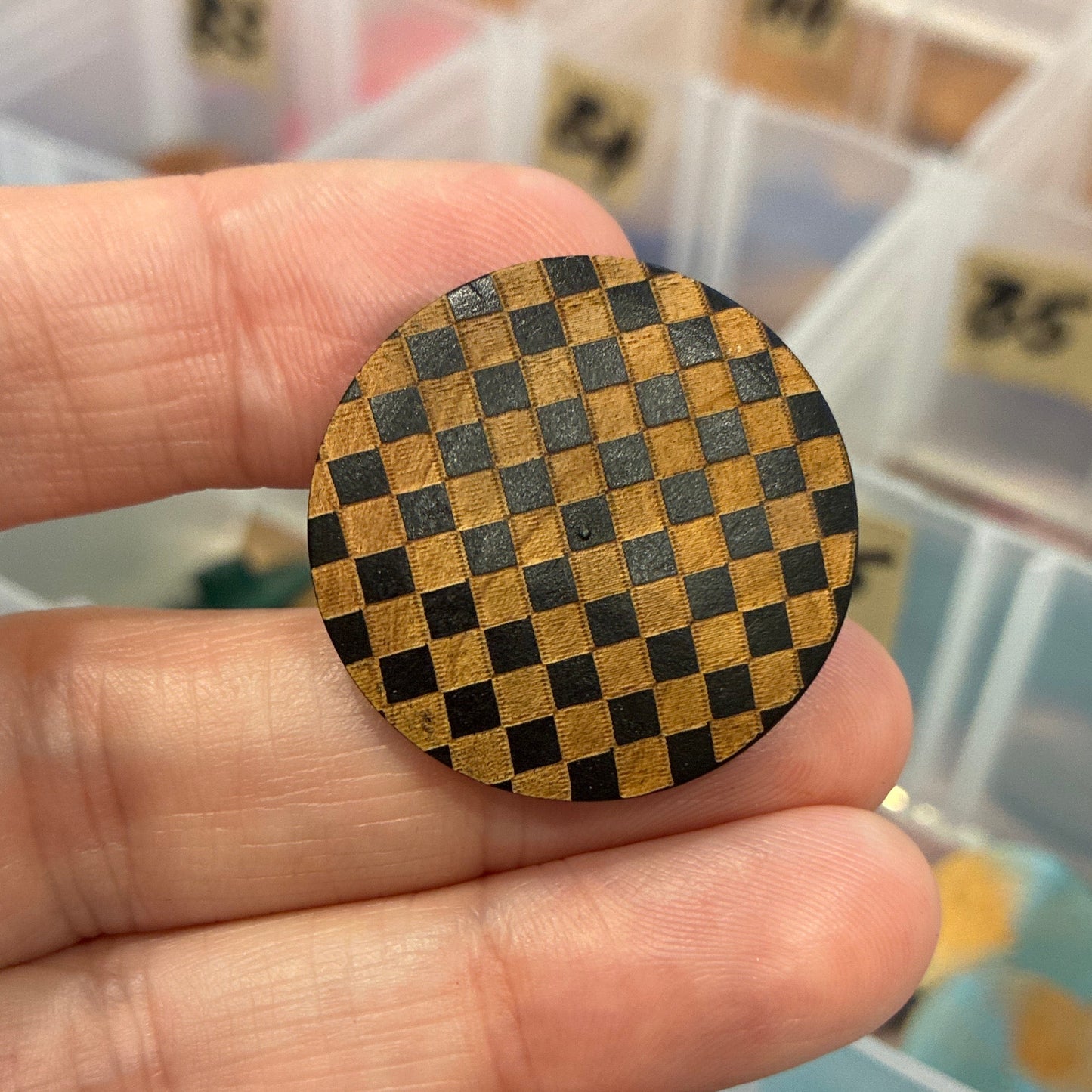 Hand holding a round wooden button with a checkered pattern in front of a container with various items.