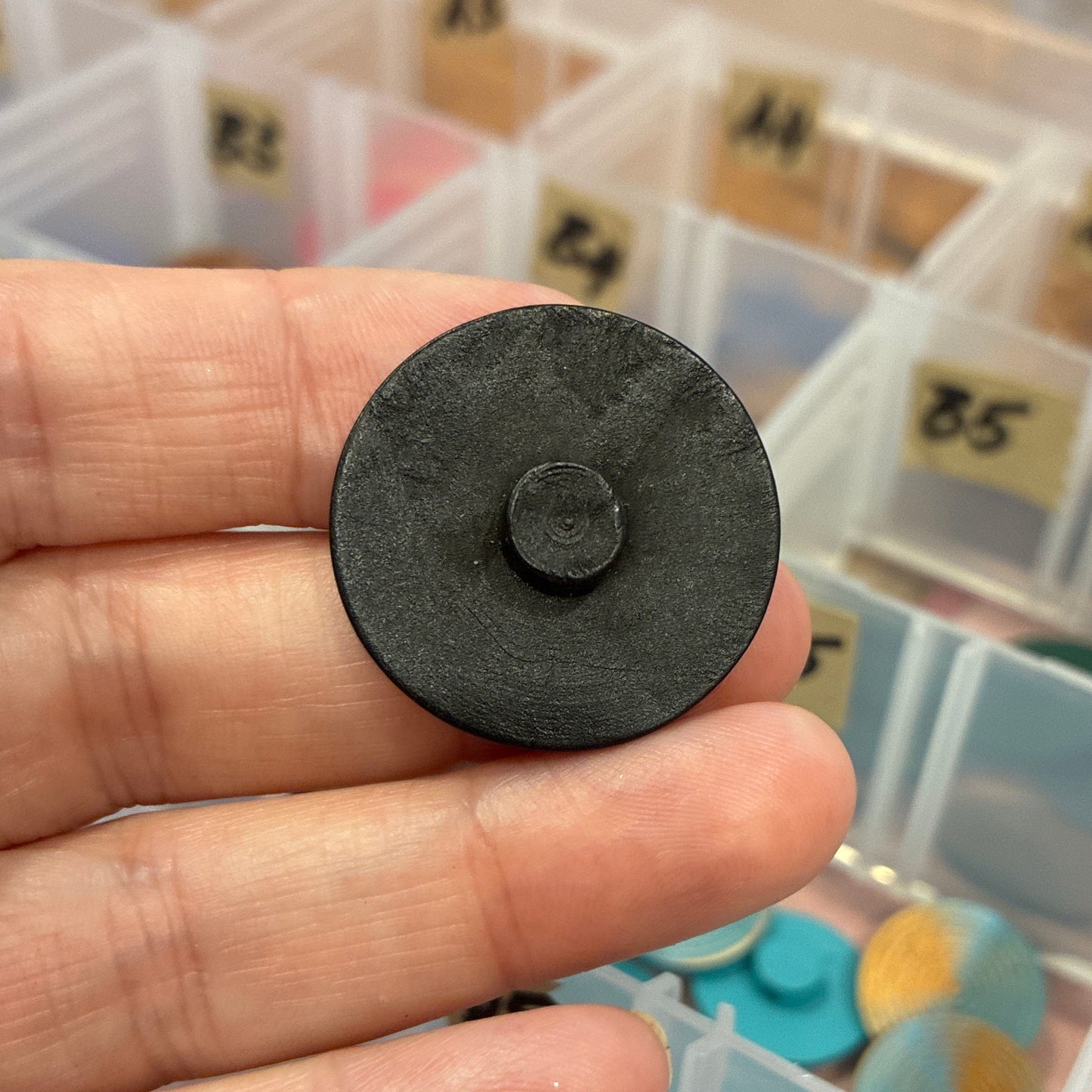 Black circular button held in a hand with a container of colorful objects in the background