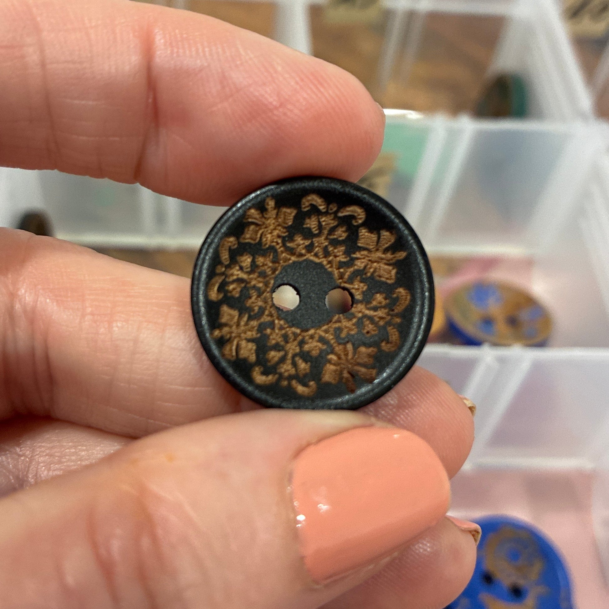 Hand holding a decorative button with intricate designs, blurred background of a storage box with more buttons.
