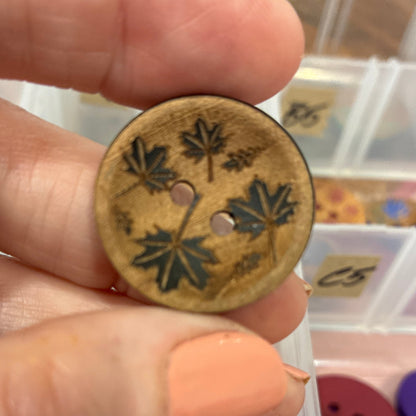 Hand holding a decorative button with floral patterns against a background of a button collection container.
