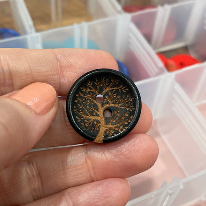 Hand holding a decorative button with intricate design in front of a container with other buttons.