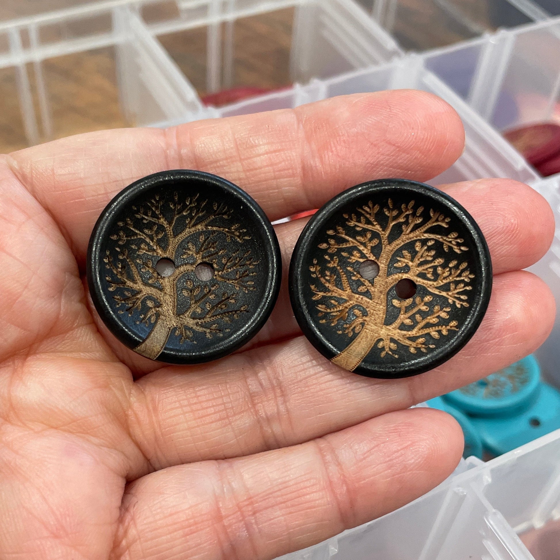 Two black buttons with tree designs held in a hand, with a container of colorful buttons in the background.