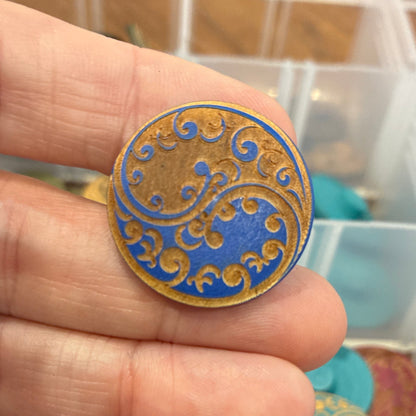 Close-up of a hand holding a decorative button with intricate gold and blue design, against a background of colorful buttons in a container.