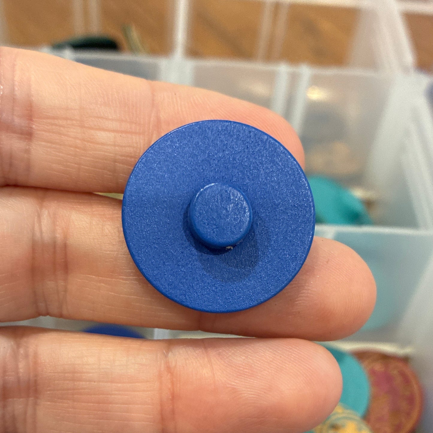 Blue circular button held between fingers with a container of colorful objects in the background