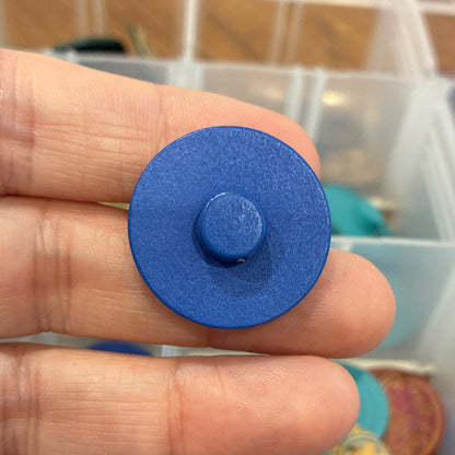 Blue circular button held between fingers with a container of colorful objects in the background