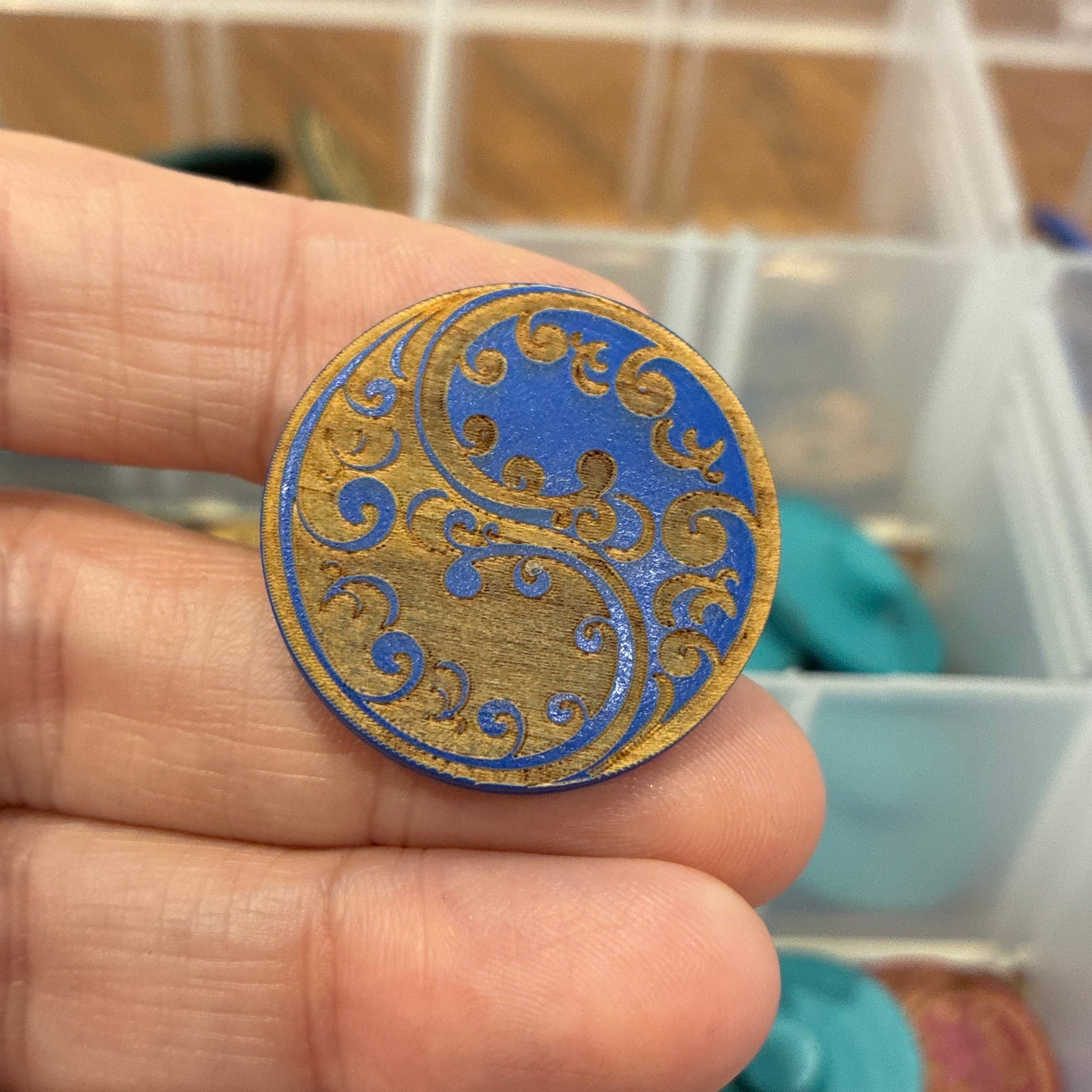 Decorative blue and gold button held between fingers with a container of similar buttons in the background.