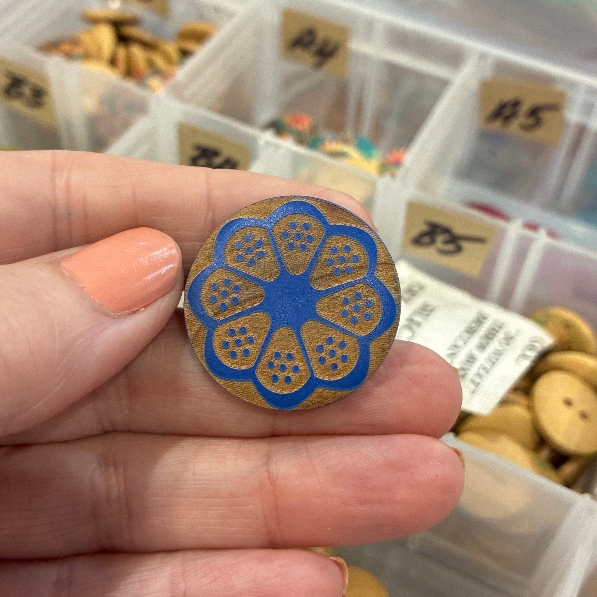 Hand holding a blue and gold decorative button with a container of buttons in the background