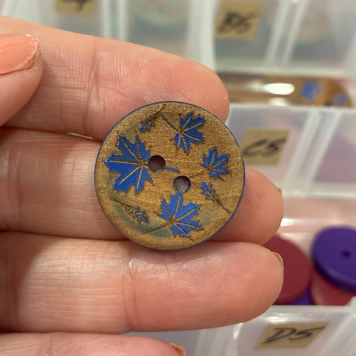Hand holding a decorative button with floral design in front of a container of buttons.
