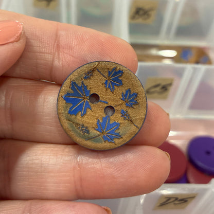 Hand holding a decorative button with floral design in front of a container of buttons.