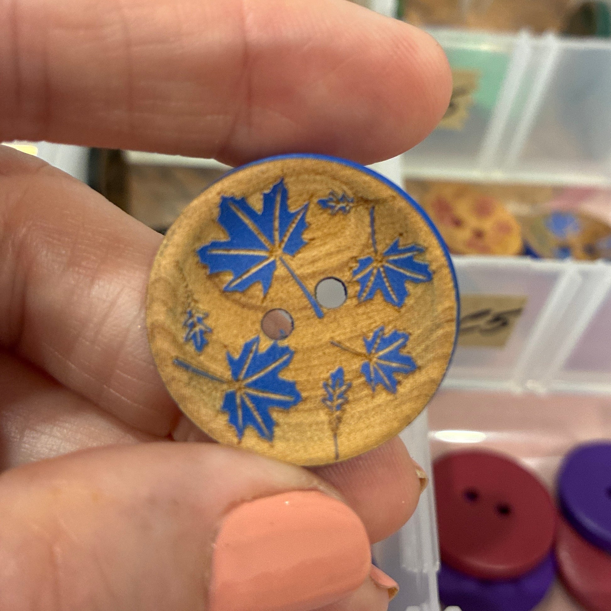 Hand holding a wooden button with blue floral design in front of a container of buttons.