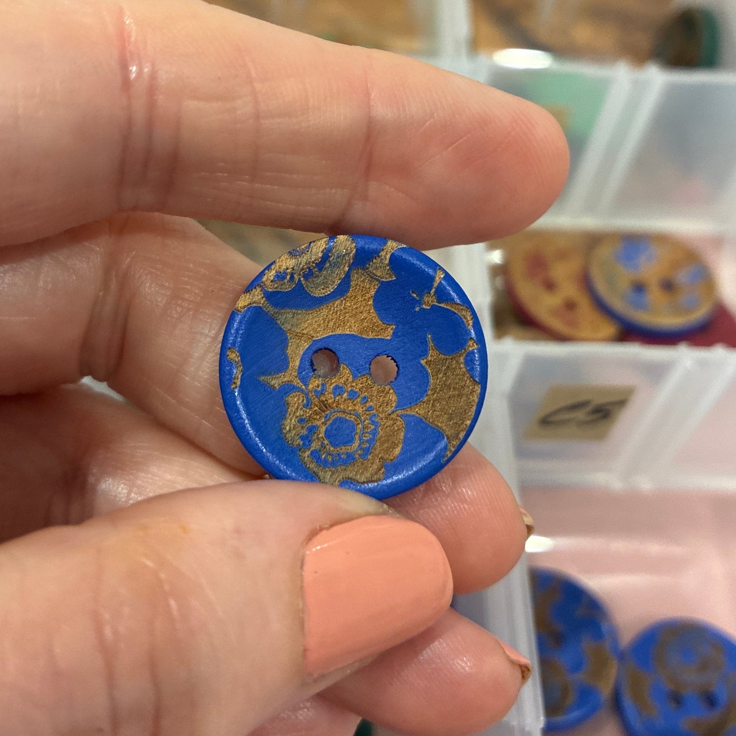 Hand holding a blue and gold decorative button with a blurred background of a storage box.