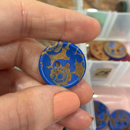 Hand holding a blue and gold decorative button with a blurred background of a storage box.