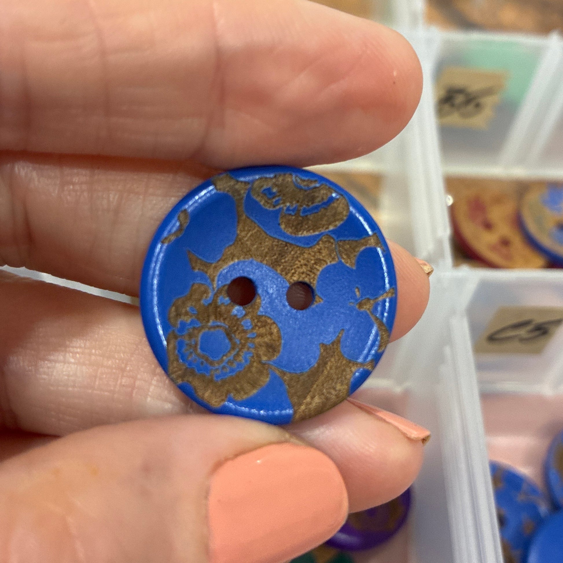 Hand holding a blue decorative button with intricate designs, in front of a clear container with more buttons.