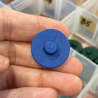 Hand holding a blue circular button with a container of buttons in the background